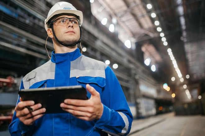 Industrial worker in safety gear and reflective jacket holding tablet in manufacturing facility.